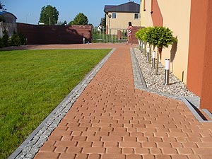 Czerwona nawierzchnia ułożona z kostki brukowej prowadząca przez trawnik i rabatę z ozdobnymi krzewami w kierunku domu z ceglanym ogrodzeniem w tle.