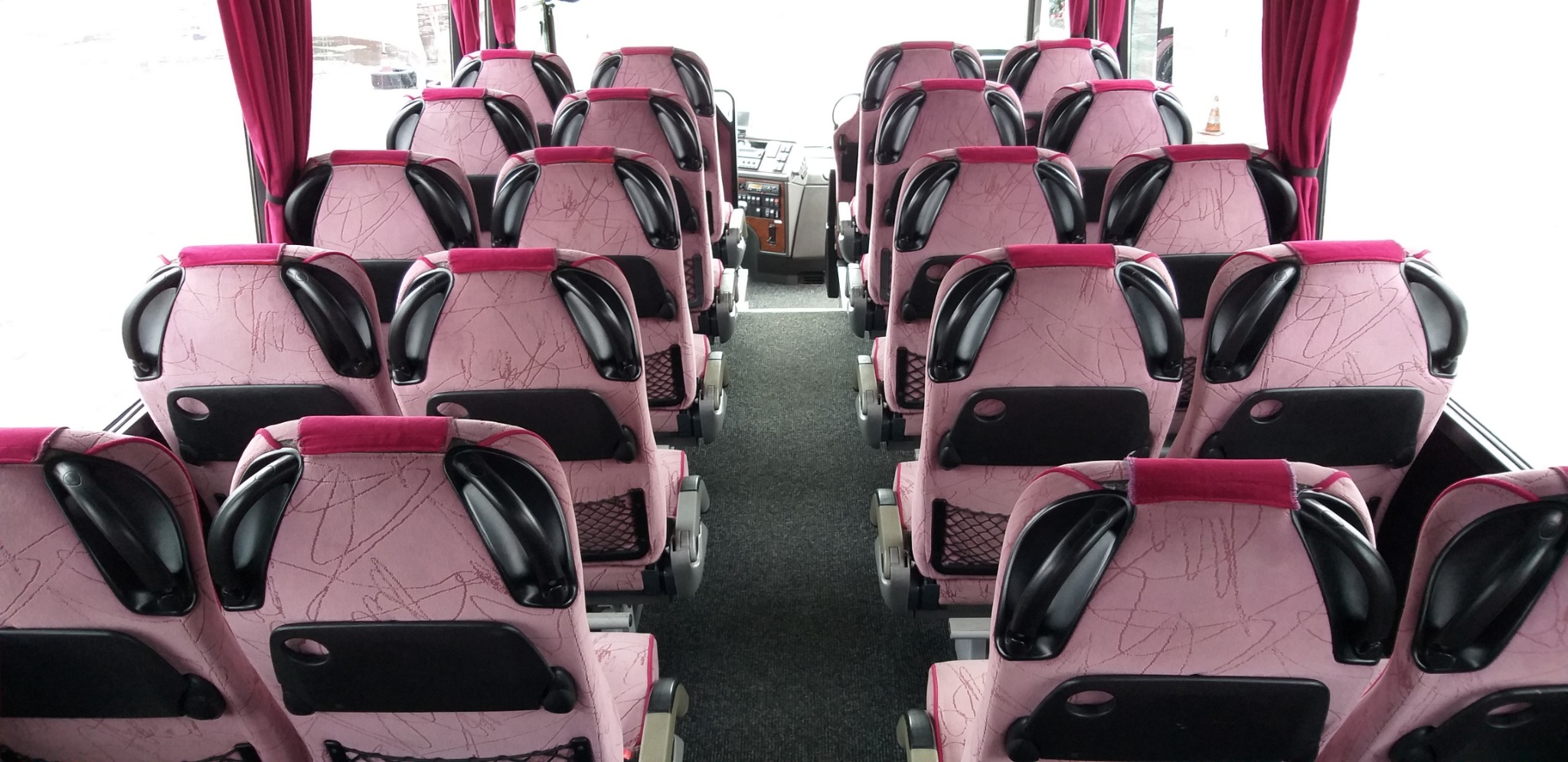 Wnętrze autobusu z rzędami różowych foteli z czarnymi zagłówkami i siatkami na oparciach, widok z przodu kabiny kierowcy.