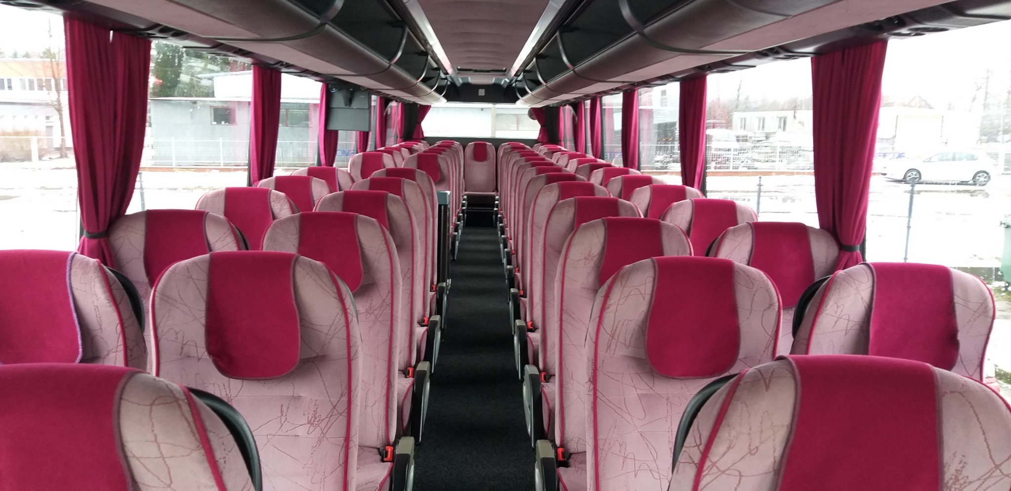 Wnętrze autobusu turystycznego z rzędami foteli w odcieniach różu i bordowych zasłon, widok z przodu kabiny.