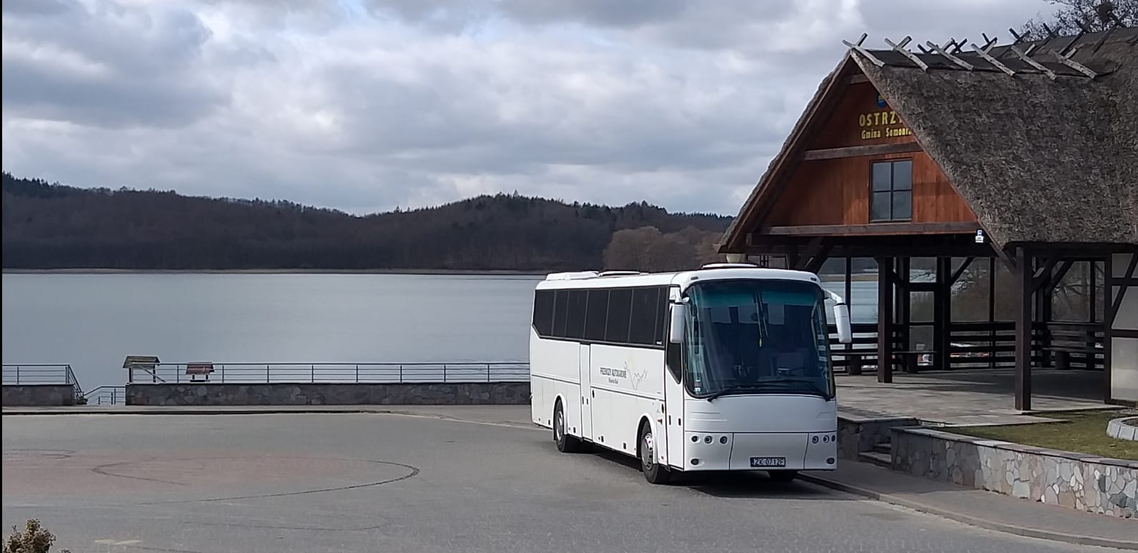 Biały autokar turystyczny zaparkowany obok drewnianej wiaty z trzcinowym dachem, w tle jezioro i zalesione wzgórza.