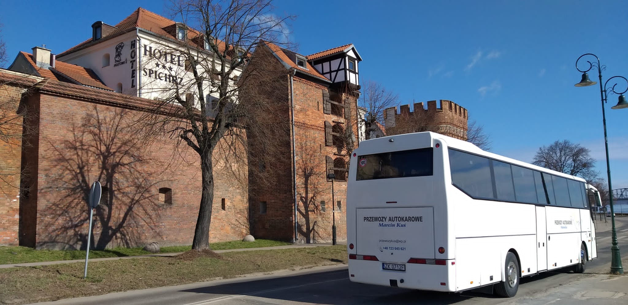 Biały autokar firmy przewozowej z Koszalina zaparkowany na tle ceglanych budynków i Hotelu Spichrz w słoneczny dzień.