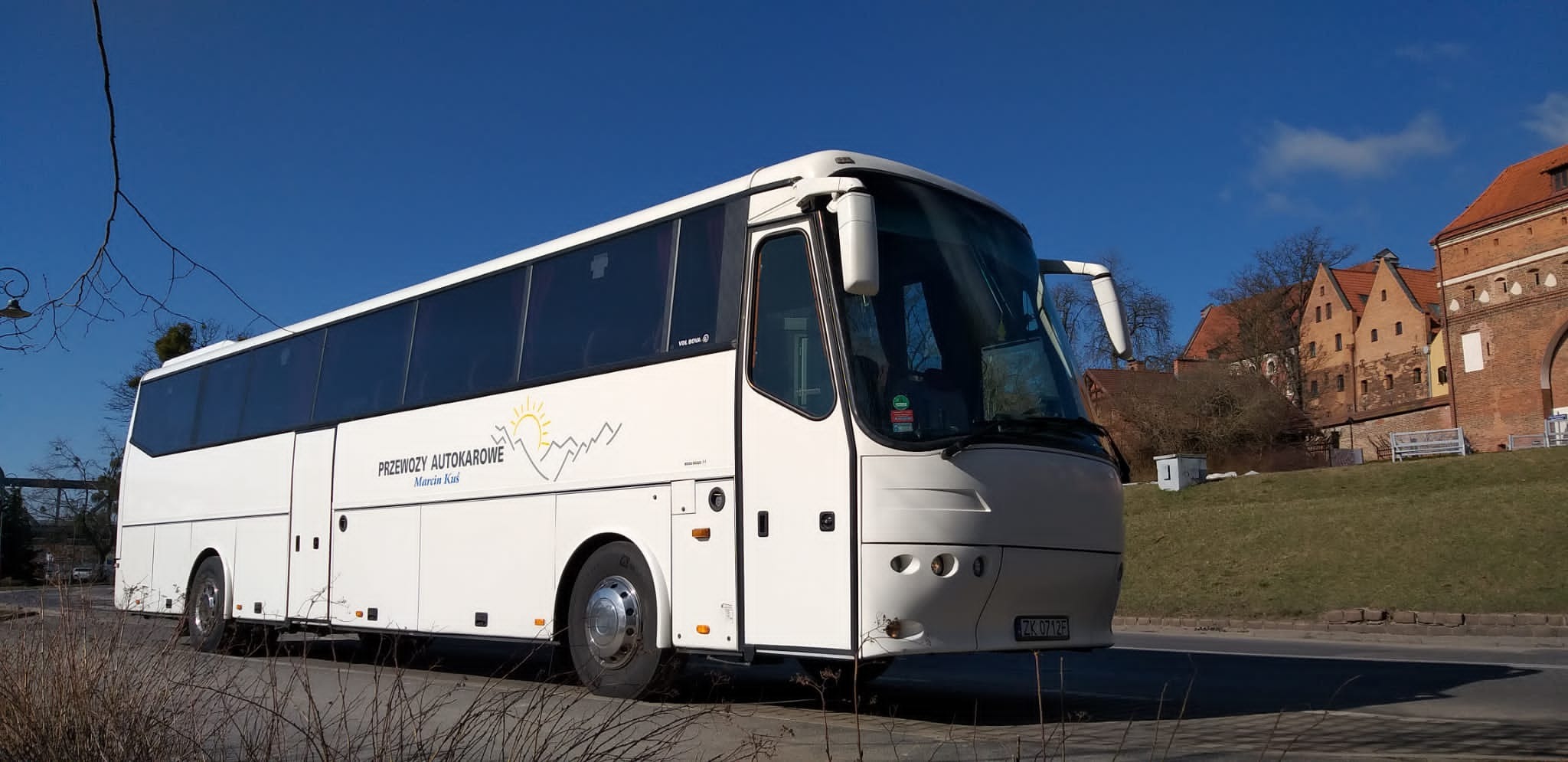 Biały autokar z logo firmy na tle ceglanego budynku i błękitnego nieba.