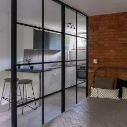 Loftowa szklana ścianka działowa z czarnymi szprosami oddzielająca sypialnię od kuchni, widoczna ceglana ściana i fragment łóżka