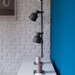 LUX GROUP SPÓŁKA Z O.O. - Czarna, regulowana lampa podłogowa o trzech kloszach oświetla trzy świece na białym blacie, na tle białej ściany z cegły i niebieskiej ściany.