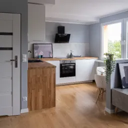 Jasna kuchnia z drewnianymi akcentami: blat z litego drewna, białe szafki, heksagonalne płytki na ścianie, okno z widokiem na zieleń, nowoczesne oświetlenie i minimalistyczne dodatki.