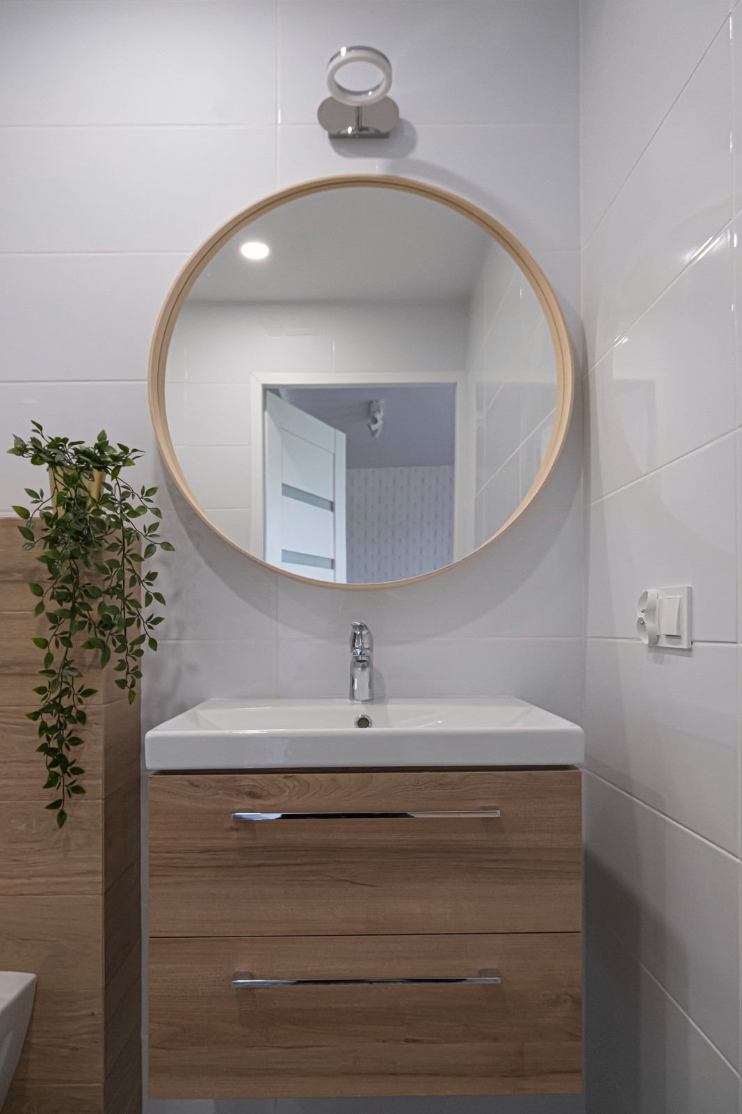 Nowoczesna łazienka z okrągłym lustrem w drewnianej ramie nad umywalką z szafką w kolorze drewna, białe płytki na ścianach i dekoracyjna roślina zwisająca z lewej strony, widoczne gniazdko...