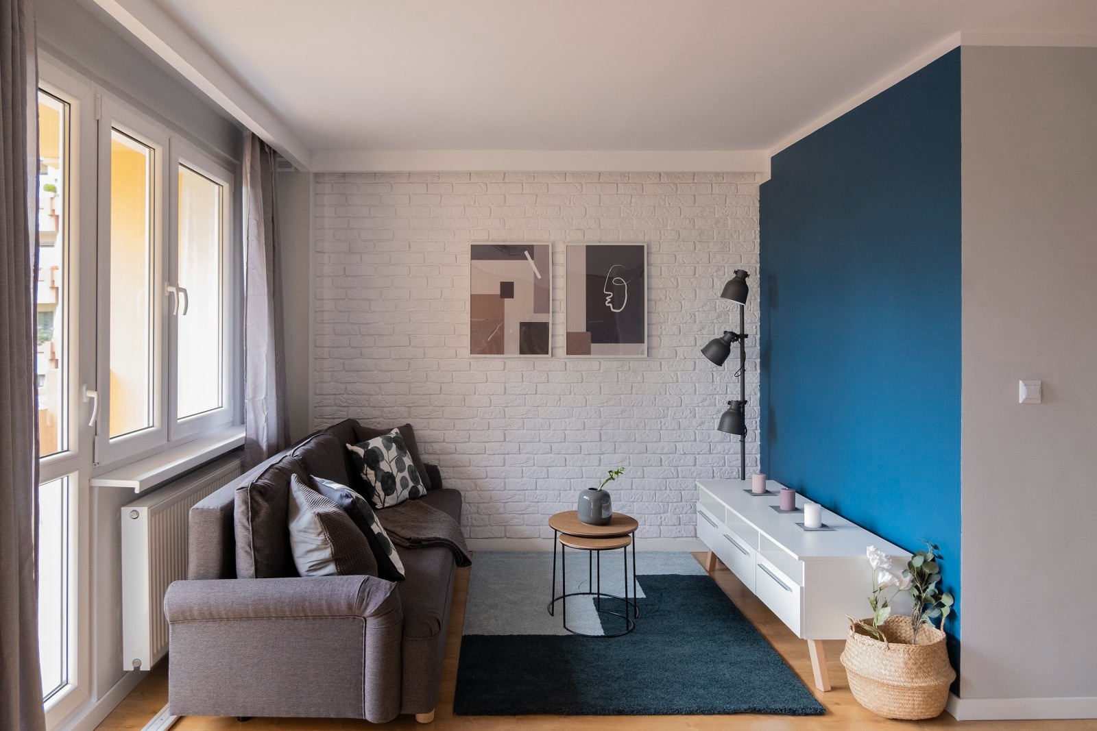 Jasne wnętrze salonu z szarą sofą, białymi oknami i ścianą z białej cegły, urozmaicone niebieską ścianą i nowoczesnymi plakatami.