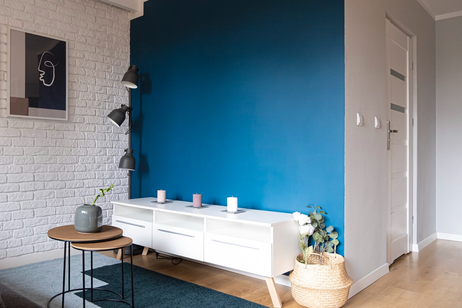 Nowoczesny salon z białą cegłą, ścianą w kolorze głębokiego błękitu i białym meblem RTV. Trzy świece i kwiaty dodają przytulności. Widoczny fragment instalacji oświetleniowej w postaci lampy...