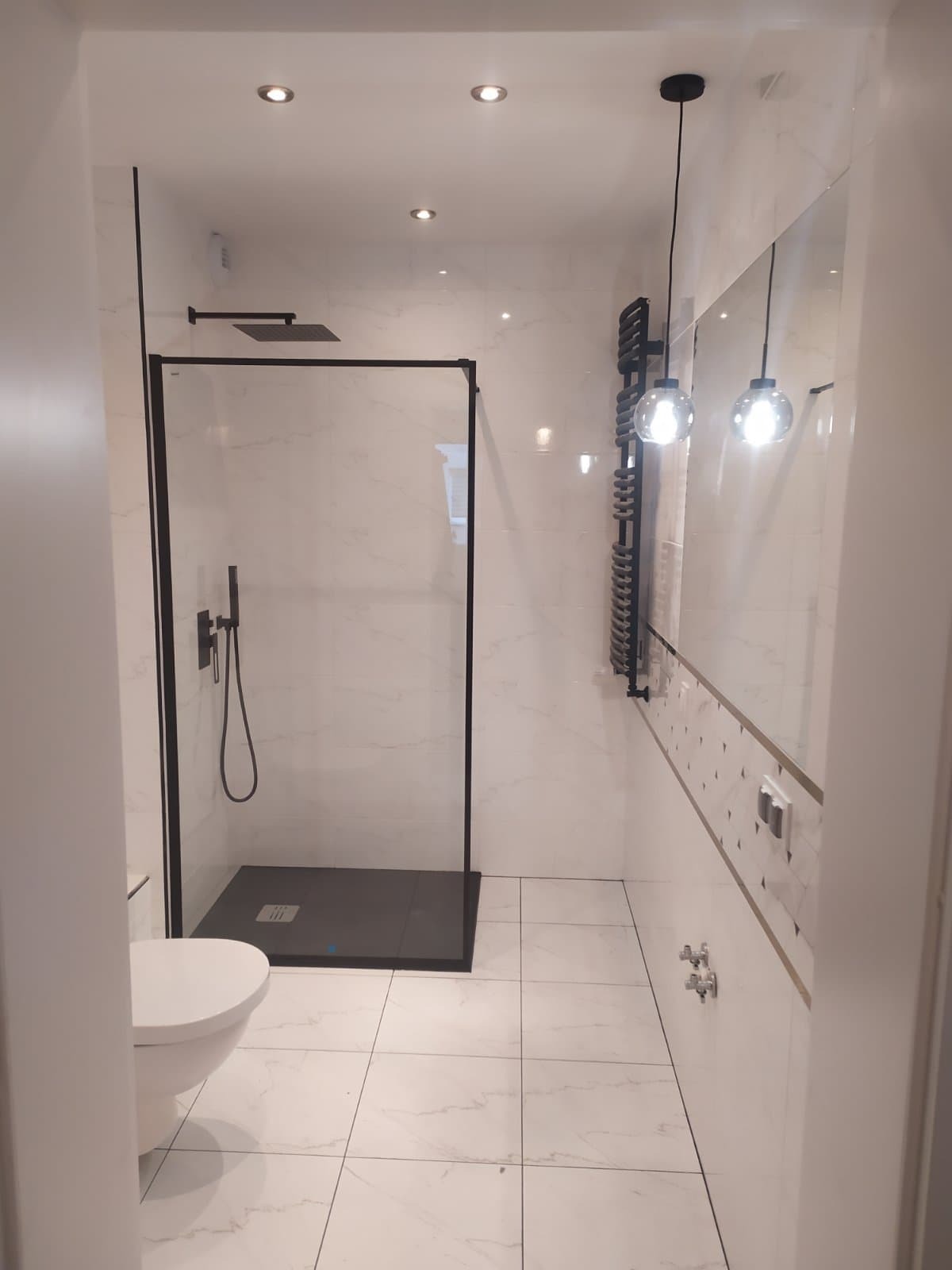 Nowoczesna łazienka z kabiną prysznicową typu walk-in z czarną ramą, grzejnikiem drabinkowym i wiszącymi lampami kulami.
