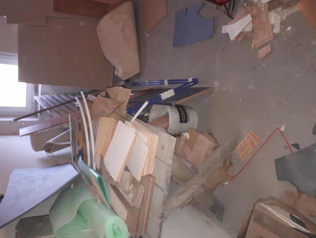 Stos gruzu i odpadów budowlanych: fragmenty płytek, kartonów, rur PCV i rolka zielonej pianki na szarym, betonowym podłożu, widoczny fragment okna w tle.