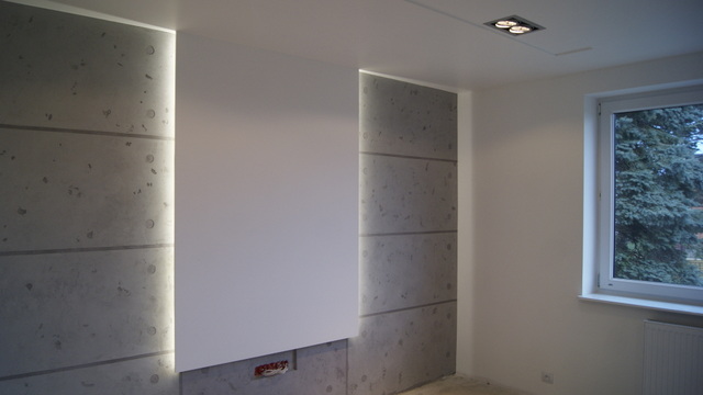 Nowoczesne wnętrze z dekoracyjnymi tynkami imitującymi beton, podświetlonymi listwami LED po bokach białej ściany, widoczne gniazdko elektryczne w ścianie i okno z widokiem na drzewo