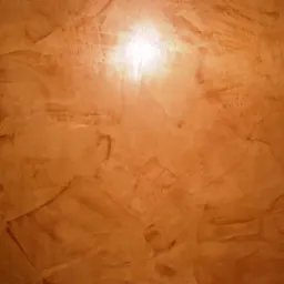 Pomarańczowa ściana z dekoracyjną fakturą imitującą kamień, oświetlona od góry, z widocznymi smugami i nierównościami w strukturze.