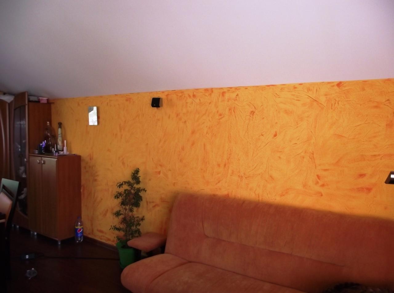 Fragment pokoju z pomarańczową ścianą o nieregularnej fakturze, brązową szafką z wyposażeniem, rośliną doniczkową i różową sofą w tle jasnego sufitu.