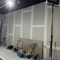 Wnętrze w trakcie montażu płyt karton-gips, widoczne spoiny wypełnione masą szpachlową, narzędzia budowlane i oświetlenie robocze.