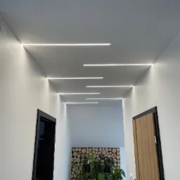 Korytarz z białymi ścianami i sufitem, oświetlony liniowymi lampami LED wpuszczonymi w sufit, widoczne drzwi w czarnej ramie i drzwi z okleiną drewnopodobną, w tle dekoracja ścienna z drewnianych...