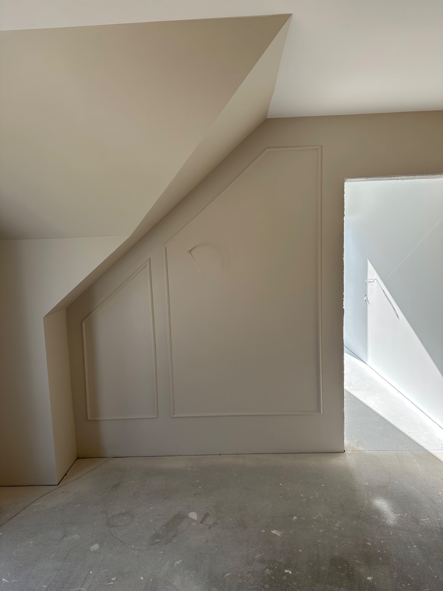 Jasne wnętrze poddasza ze ścianami w odcieniach beżu, dekoracyjnymi listwami i surową podłogą. Widoczne niedoskonałości na ścianie i brak wykończenia.