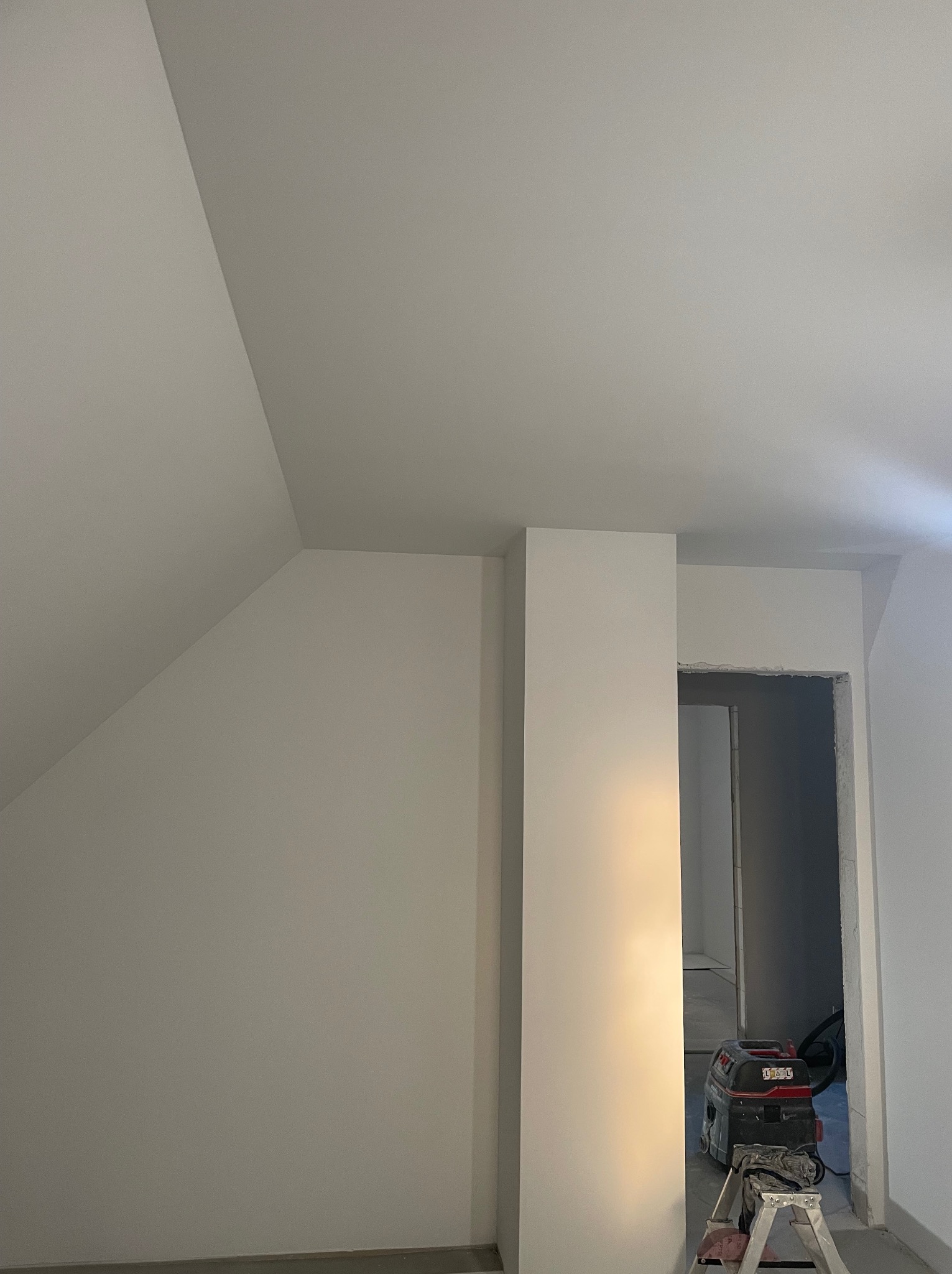 Wnętrze po remoncie: białe ściany i sufit na poddaszu, widoczny fragment futryny drzwi i odkurzacz przemysłowy na drabinie. Minimalistyczny design, świeżo po malowaniu.