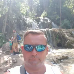 Mężczyzna w okularach przeciwsłonecznych robiący selfie na tle malowniczego wodospadu i bujnej zieleni. W odbiciu okularów widoczne otoczenie.