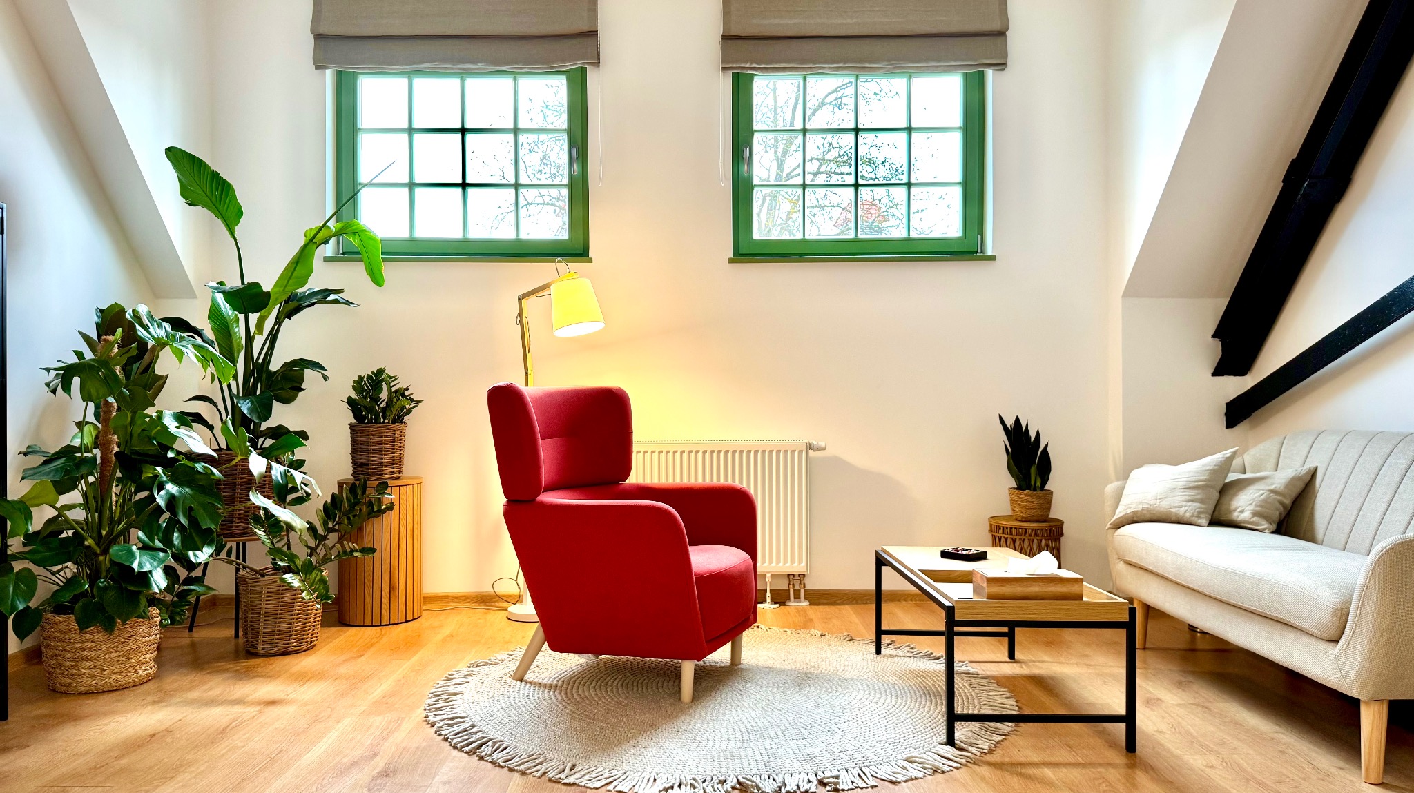 Jasne wnętrze pokoju z czerwonym fotelem, beżową sofą, drewnianym stolikiem kawowym i dużą ilością roślin doniczkowych, oświetlone lampą podłogową i naturalnym światłem z okien z zielonymi ramami.