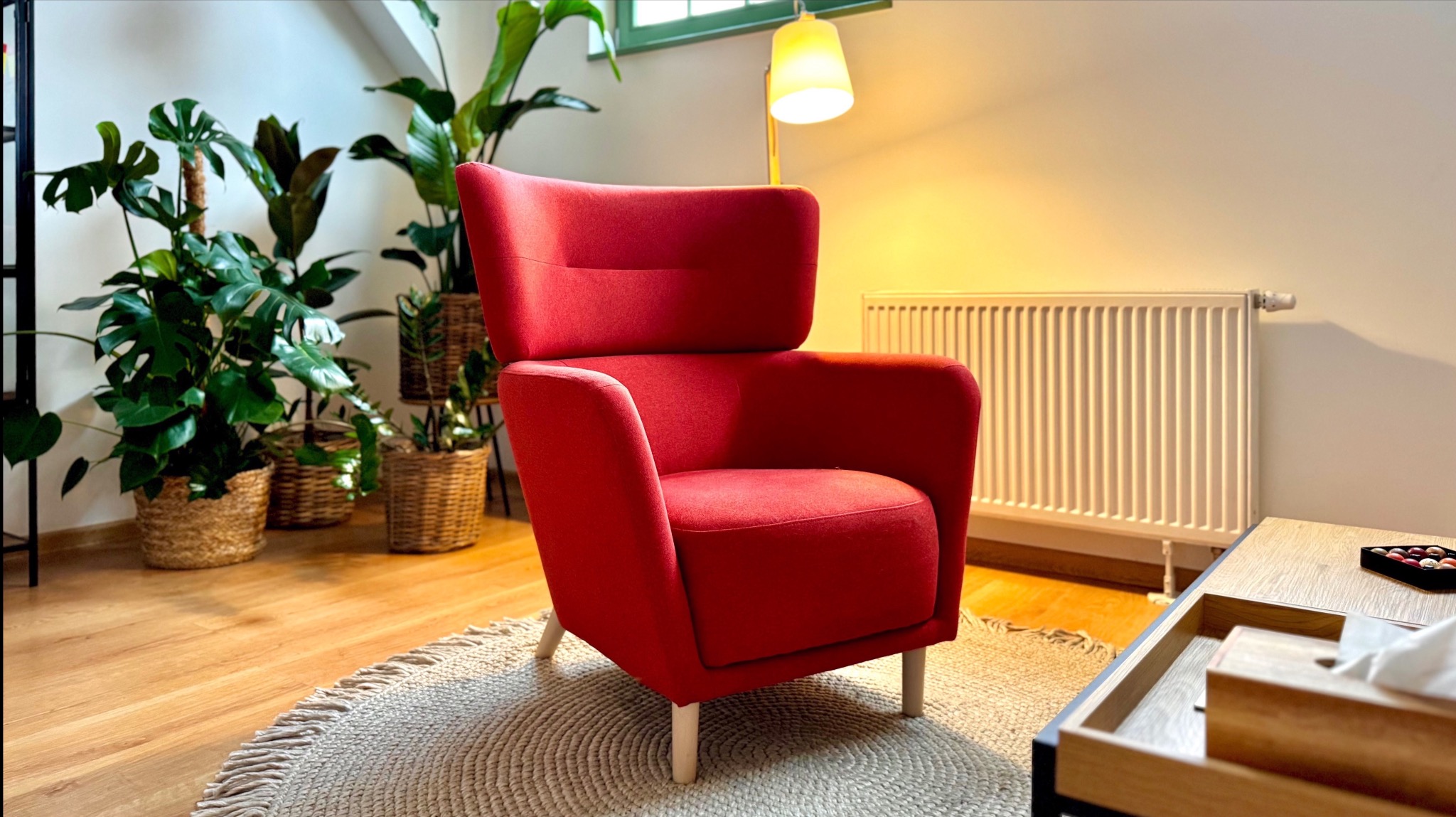Czerwony fotel z wysokim oparciem na okrągłym dywaniku w jasnym gabinecie, obok roślin doniczkowych i grzejnika.