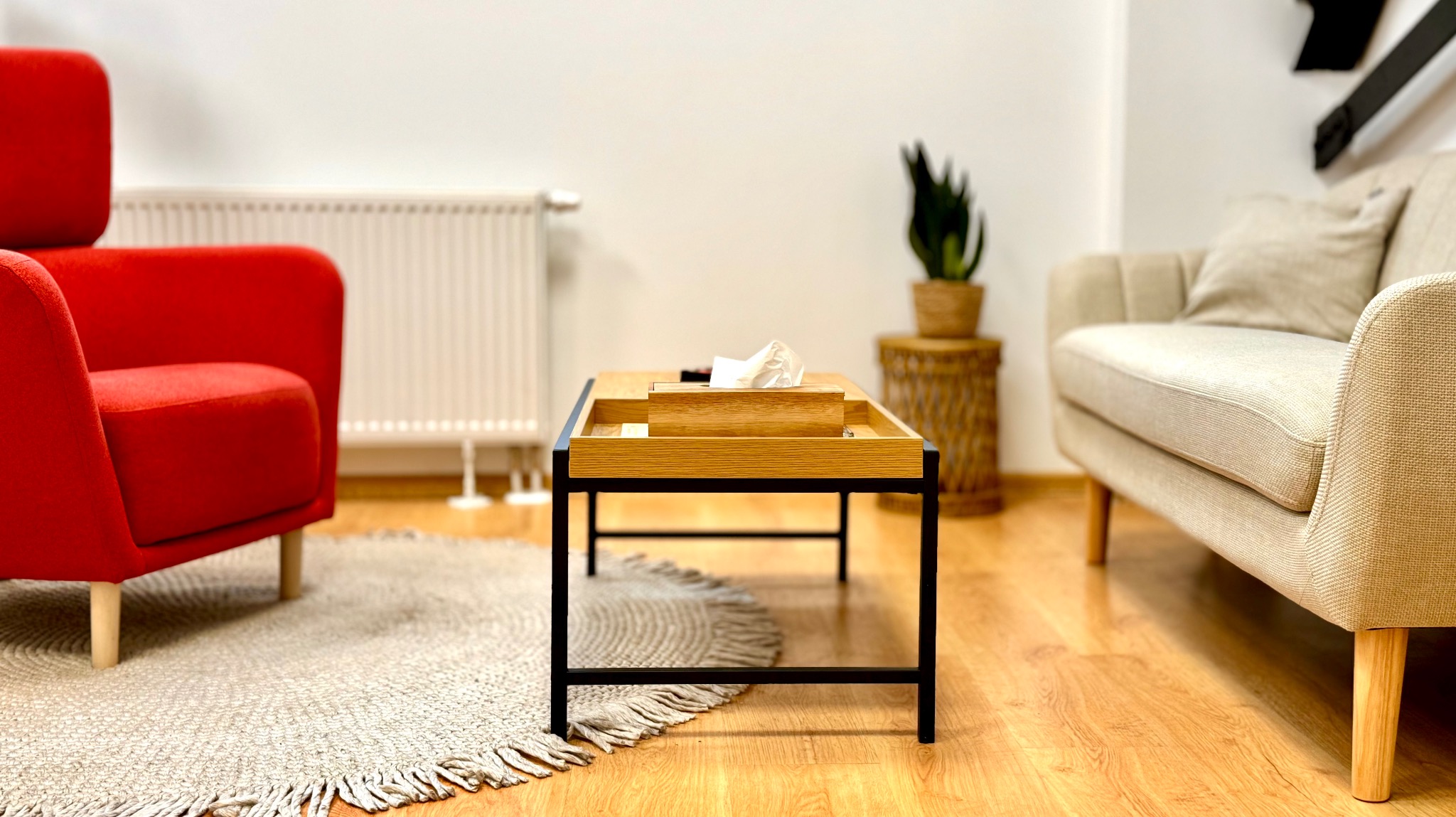 Jasne wnętrze gabinetu z czerwonym fotelem, beżową sofą, drewnianym stolikiem kawowym z pudełkiem chusteczek, okrągłym dywanem i rośliną doniczkową.
