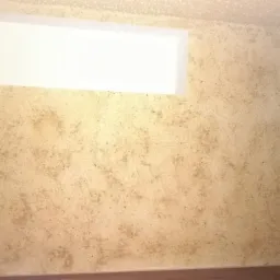 Ściana wewnętrzna z nałożoną gładzią dekoracyjną w odcieniach beżu i brązu, z widocznym oknem i fragmentem drewnianej framugi drzwi.