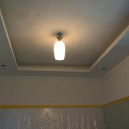 Cis-Bud - Pomieszczenie w trakcie prac wykończeniowych, zabezpieczone folią i taśmą malarską, z widocznym sufitem podwieszanym z oświetleniem punktowym i centralną lampą.