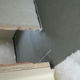 Cis-Bud - Przygotowanie podłoża pod gładzie: surowy tynk cementowo-wapienny na ścianie, widoczne fragmenty cegieł i wystające przewody elektryczne.