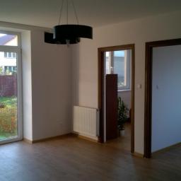 Cis-Bud - Jasne wnętrze pokoju z dużym oknem wychodzącym na ogród, drewnianą podłogą, białymi ścianami, nowym grzejnikiem i czarną, nowoczesną lampą sufitową