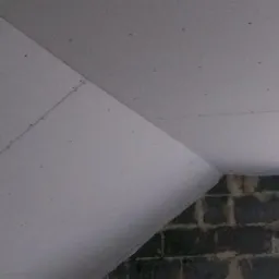 Skośny sufit na poddaszu, częściowo pokryty szarą płytą, widoczne łączenia płyt i wkręty, fragment ściany z szarego betonu komórkowego i okno w tle.