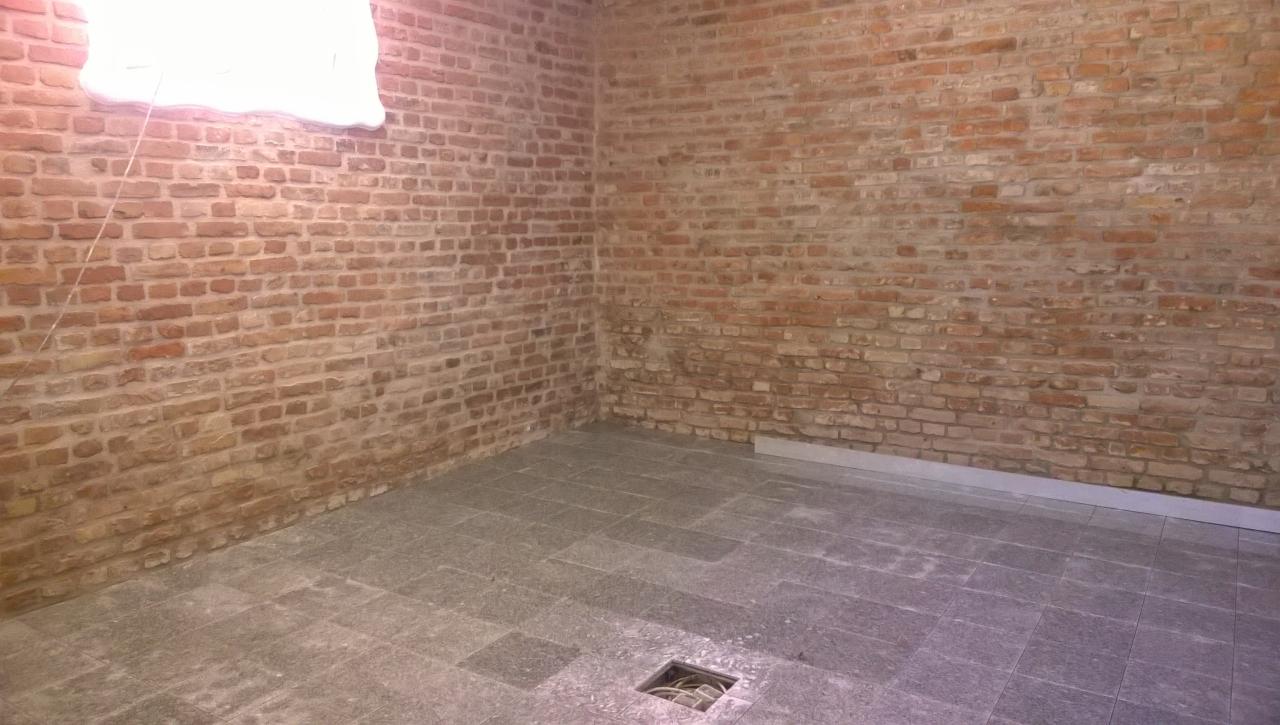 Surowa przestrzeń z ceglanymi ścianami i szarą kamienną podłogą, widoczny otwór kanalizacyjny i listwa przypodłogowa