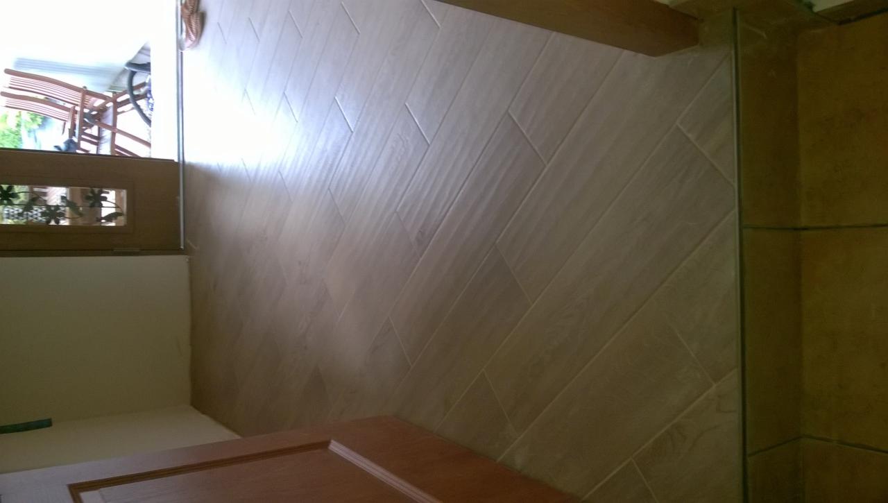 Ściana wyłożona podłużnymi, beżowymi płytkami ceramicznymi imitującymi drewno, ułożonymi w jodełkę, widok z bliska na narożnik pomieszczenia.