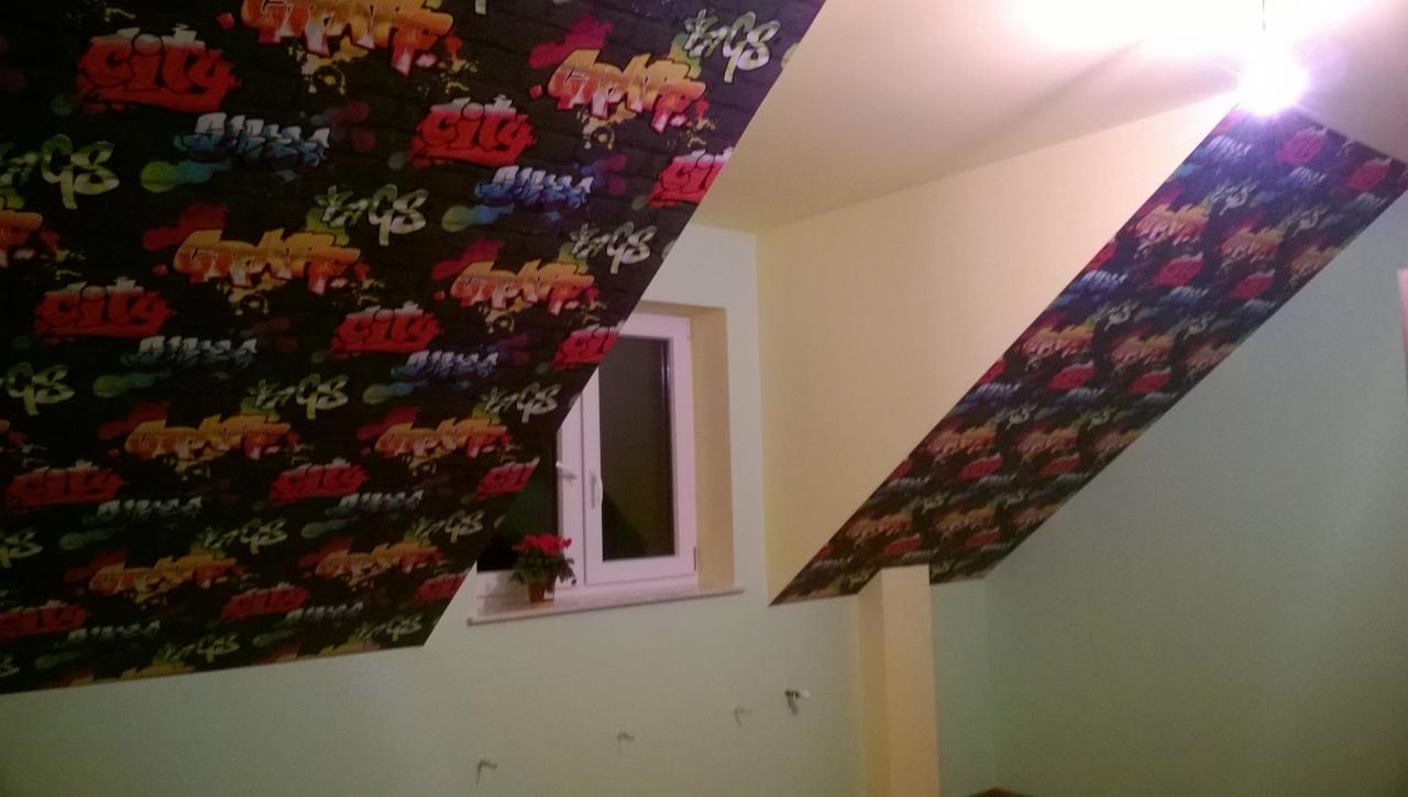 Poddasze po remoncie z ukośnymi ścianami wykończonymi tapetą w stylu graffiti, okno z kwiatami na parapecie, ściany pomalowane na jasnozielony kolor.