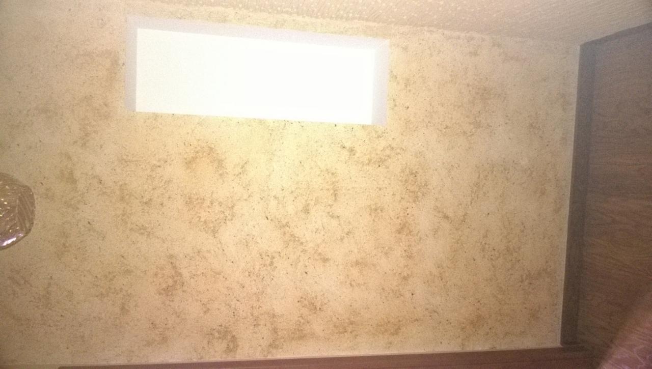 Ściana wewnętrzna z nałożoną gładzią dekoracyjną w odcieniach beżu i brązu, z widocznym oknem i fragmentem drewnianej framugi drzwi.