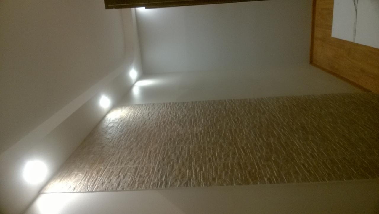 Ściana wykończona dekoracyjną, teksturowaną okładziną w odcieniach beżu, oświetlona punktowymi lampami LED w suficie pod skosem, z fragmentem obrazu w drewnianej ramie w prawym górnym rogu.