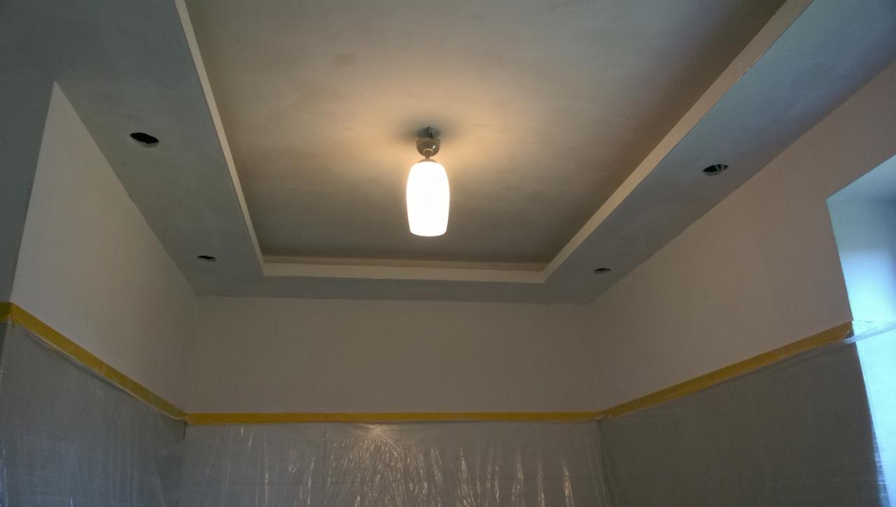 Pomieszczenie w trakcie prac wykończeniowych, zabezpieczone folią i taśmą malarską, z widocznym sufitem podwieszanym z oświetleniem punktowym i centralną lampą.
