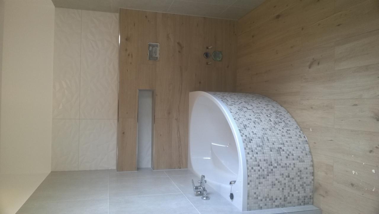 Nowoczesna łazienka z wanną obudowaną mozaiką, drewnopodobnymi panelami ściennymi i płytkami o trójwymiarowej strukturze na ścianie.