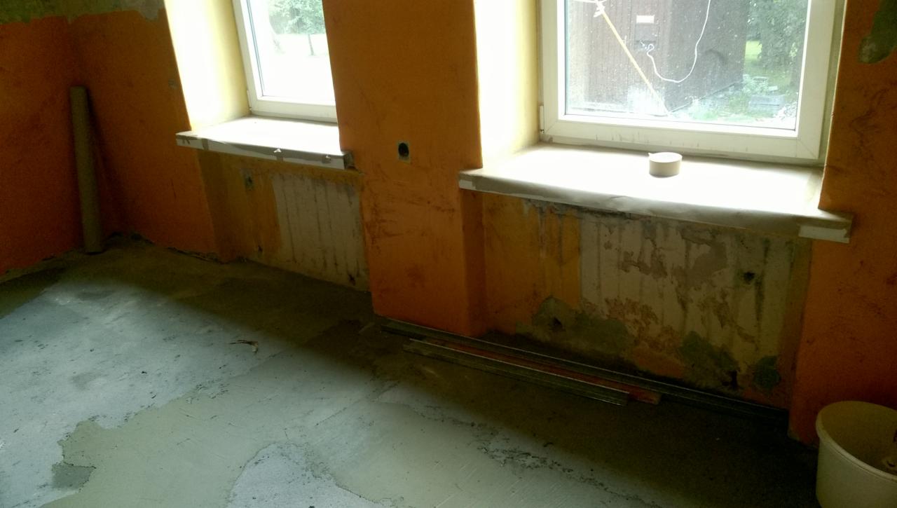 Pomarańczowe ściany w trakcie przygotowania do prac wykończeniowych, widoczne fragmenty starej farby, okna z parapetami zabezpieczonymi taśmą, surowy beton na podłodze.