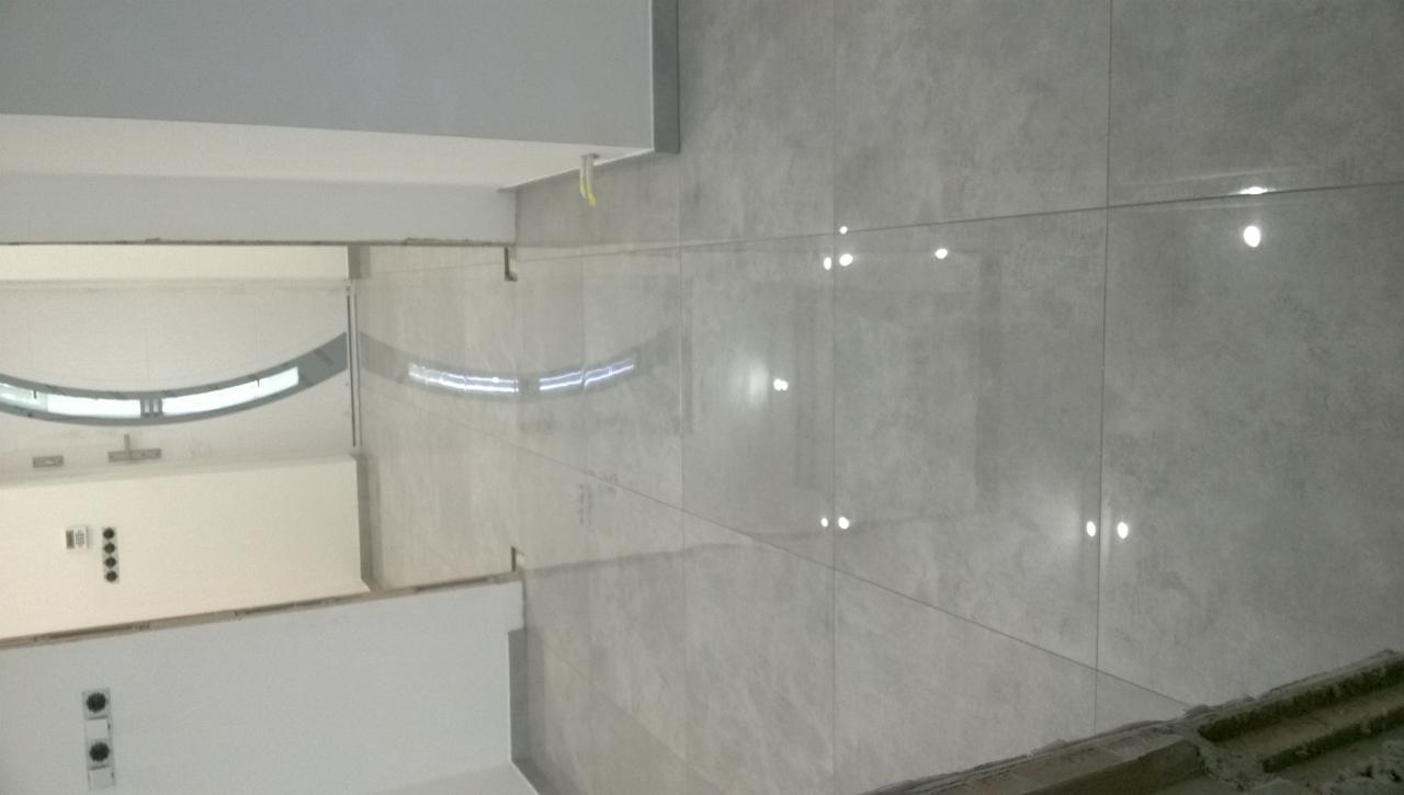 Świeżo położone, błyszczące, szare płytki na ścianie w pomieszczeniu w Mokrzyskach, z widocznym fragmentem sufitu i przygotowaną podłogą.