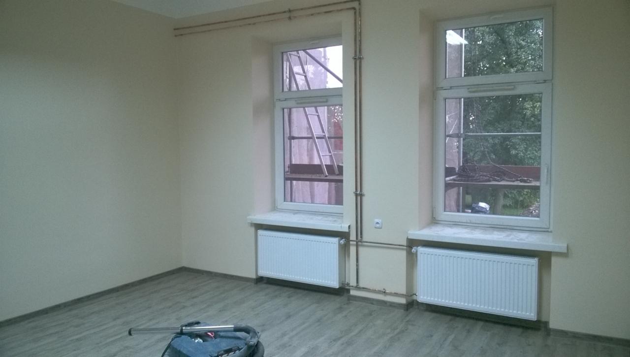 Pokój po remoncie: świeżo pomalowane ściany w odcieniach beżu, dwa okna z białymi ramami, pod którymi zamontowano białe grzejniki, na podłodze panele imitujące drewno, widoczny sprzęt malarski w rogu.