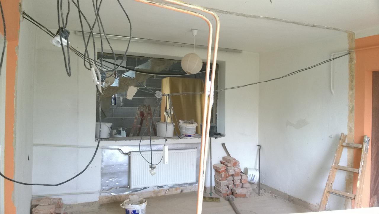 Wnętrze w trakcie remontu: odsłonięte cegły, wiszące przewody elektryczne, grzejnik, wiadro z farbą, drabina oparta o ścianę, widoczna lampa w kształcie kuli.