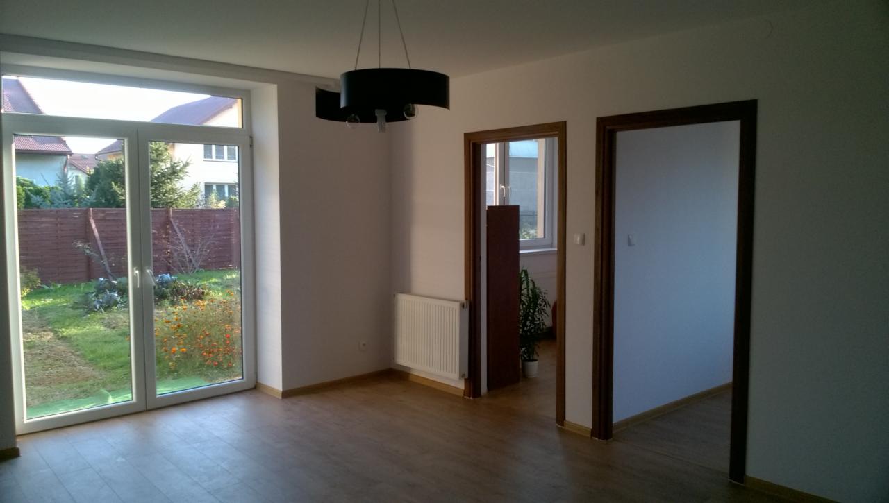 Jasne wnętrze pokoju z dużym oknem wychodzącym na ogród, drewnianą podłogą, białymi ścianami, nowym grzejnikiem i czarną, nowoczesną lampą sufitową