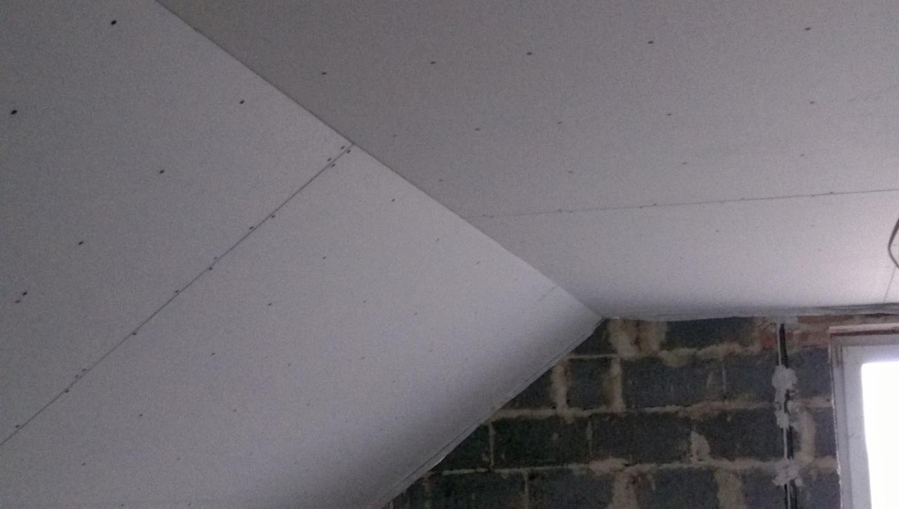 Skośny sufit na poddaszu, częściowo pokryty szarą płytą, widoczne łączenia płyt i wkręty, fragment ściany z szarego betonu komórkowego i okno w tle.