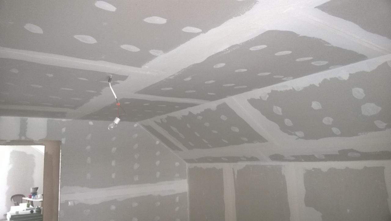 Sufit i ściany w trakcie prac wykończeniowych, widoczne płyty gipsowo-kartonowe z zaszpachlowanymi łączeniami i widocznymi wkrętami, surowa żarówka zwisająca z sufitu, pomieszczenie w stanie przed...