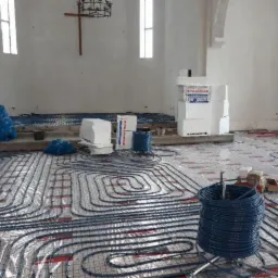 Montaż ogrzewania podłogowego w przestronnym wnętrzu, widoczne rury grzewcze ułożone na siatce, materiały izolacyjne i narzędzia, surowy wystrój wnętrza z elementami sakralnymi.