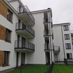 Nowoczesny budynek wielorodzinny z balkonami zabezpieczonymi ciemnymi, metalowymi balustradami, widok z poziomu trawnika oddzielonego niskim ogrodzeniem.