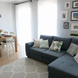 Jasne wnętrze pokoju dziennego z szarą sofą narożną, drewnianym stołem jadalnym, białymi krzesłami i dywanem w marokański wzór, oświetlone naturalnym światłem z okien.