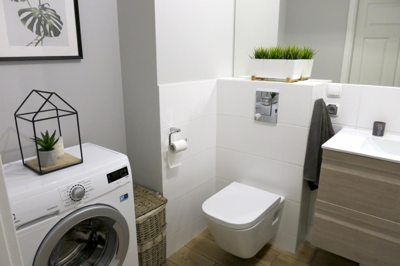 Nowoczesna łazienka z pralką, wiszącą miską WC, umywalką z szafką z drewnopodobnym fornirem, dekoracjami w postaci roślin w doniczkach i grafiki z motywem roślinnym w czarnej ramie.