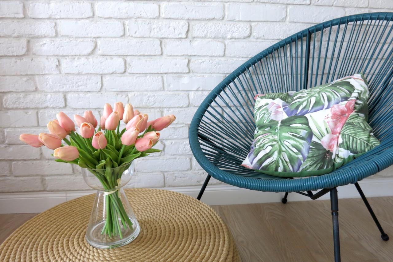 Turkusowe krzesło Acapulco z poduszką w motywy tropikalne i bukiet różowych tulipanów w szklanym wazonie na okrągłym, plecionym stoliku, na tle białej ściany z cegły.