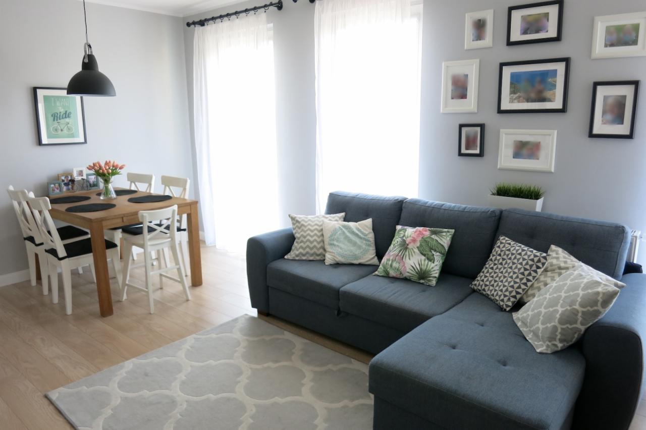 Jasne wnętrze pokoju dziennego z szarą sofą narożną, drewnianym stołem jadalnym, białymi krzesłami i dywanem w marokański wzór, oświetlone naturalnym światłem z okien.